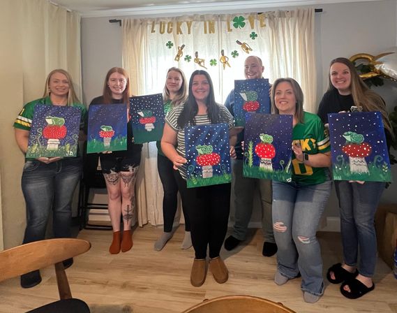 Seven friends in a cozy living room painting party, each holding a canvas of a red-spotted mushroom with green accents and starry night backgrounds under a “Lucky in Love” shamrock banner.