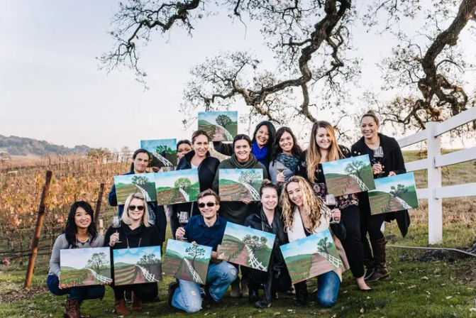 Smiling group in a vineyard holding matching landscape canvases and wine glasses at an outdoor paint-and-sip party beneath a sprawling oak and white fence