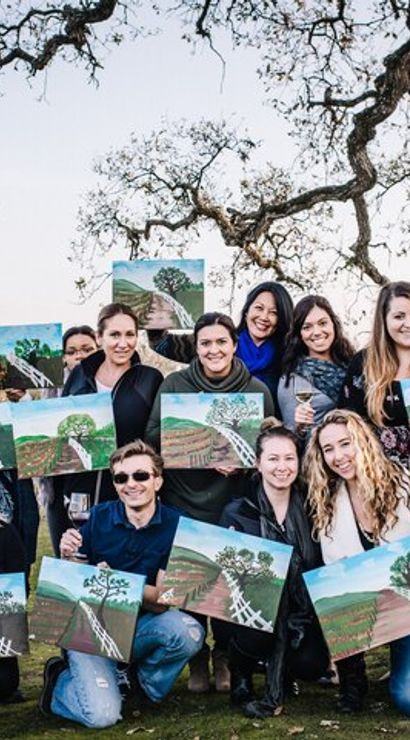Smiling group of people at a vineyard painting party, each holding a landscape canvas and wine glass under oak trees by a white fence with rolling vineyard rows