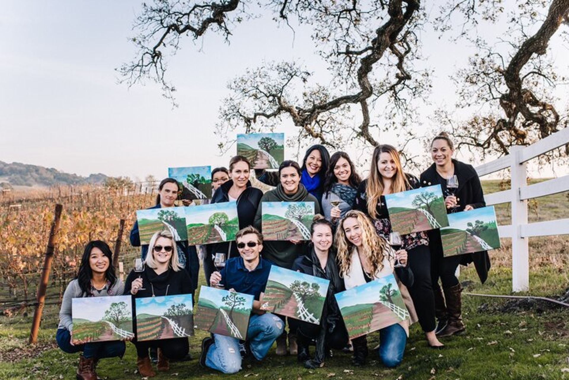 Smiling group of people at a vineyard painting party, each holding a landscape canvas and wine glass under oak trees by a white fence with rolling vineyard rows