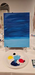 Canvas on a tabletop easel with a two-tone blue acrylic gradient suggesting sky and sea, with a paper-plate palette of yellow, red, black, navy and light-blue paints in an indoor painting class