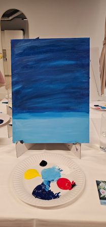 Canvas on a tabletop easel with a two-tone blue acrylic gradient suggesting sky and sea, with a paper-plate palette of yellow, red, black, navy and light-blue paints in an indoor painting class