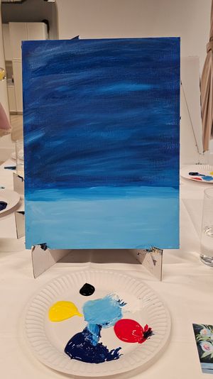 Canvas on a tabletop easel with a two-tone blue acrylic gradient suggesting sky and sea, with a paper-plate palette of yellow, red, black, navy and light-blue paints in an indoor painting class