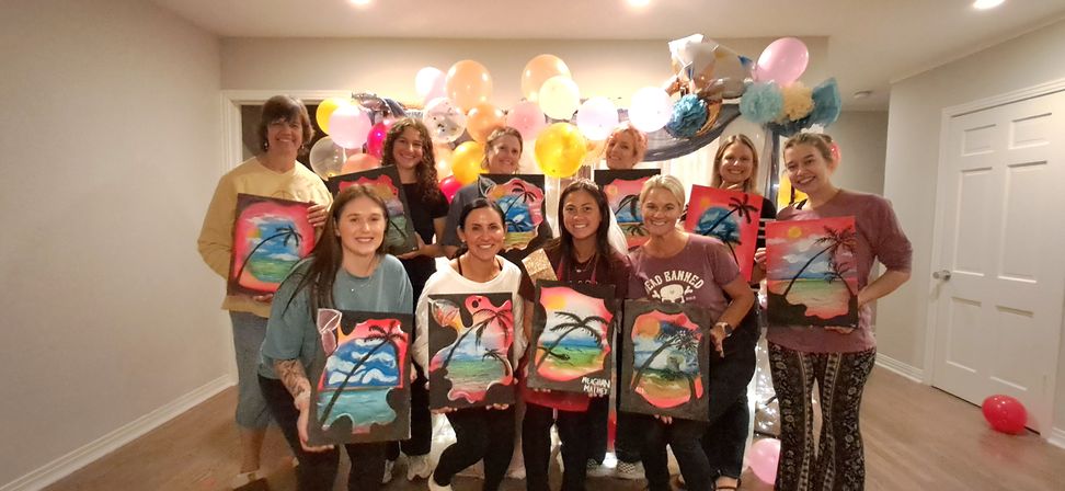 Group of women at an indoor paint‑night party, smiling and holding colorful tropical palm‑tree canvases in front of a pastel balloon backdrop