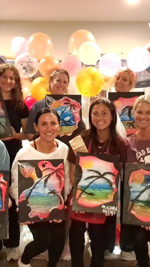 Group of women at an indoor paint‑night party, smiling and holding colorful tropical palm‑tree canvases in front of a pastel balloon backdrop