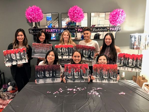 Group of women at a bachelorette painting party holding matching red-and-black tree canvases in a decorated indoor studio with pink pom-pom decorations and a party banner