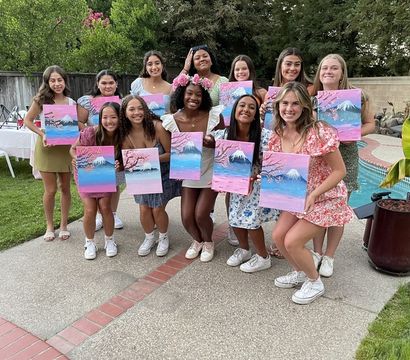 Smiling group of friends at a backyard poolside painting party, each holding a pink-and-blue canvas of a snow-capped mountain with cherry blossoms, patio, lawn and trees in the background.