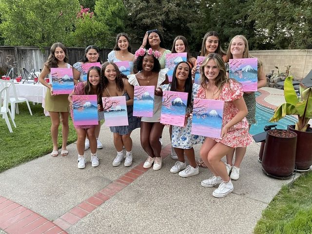Smiling group of friends at a backyard poolside painting party, each holding a pink-and-blue canvas of a snow-capped mountain with cherry blossoms, patio, lawn and trees in the background.