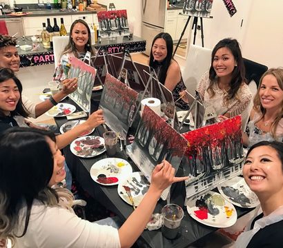 Smiling group of eight women at an indoor paint-and-sip bachelorette party around a kitchen table, painting red autumn-tree canvases with palettes and drinks