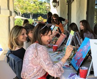 Group of women at an outdoor sip-and-paint on a sunny patio, laughing as they paint colorful floral canvases.