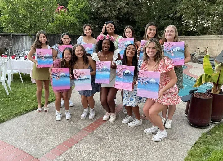 A dozen teenage girls at a backyard pool painting party, smiling and holding pink-and-blue mountain-and-cherry-blossom canvases, dressed in summer outfits and sneakers.