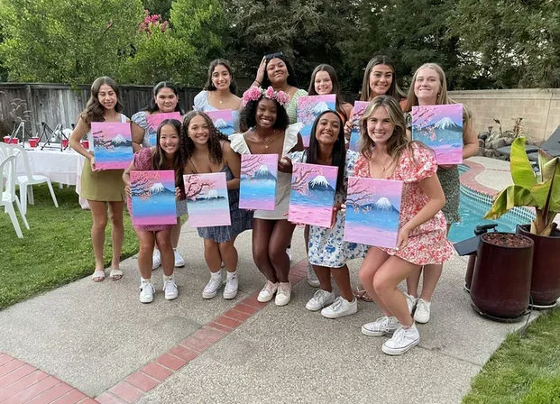 A dozen teenage girls at a backyard pool painting party, smiling and holding pink-and-blue mountain-and-cherry-blossom canvases, dressed in summer outfits and sneakers.