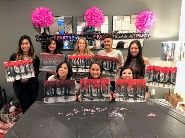 Group of women at an indoor bachelorette paint party, smiling and holding red‑leaf tree canvases under pink pom‑poms and a "Bachelorette Party" banner.