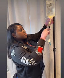 Nurse wearing pink gloves adjusts an IV drip bag on a pole in a patient room, jacket sleeve clearly labeled "NURSE".