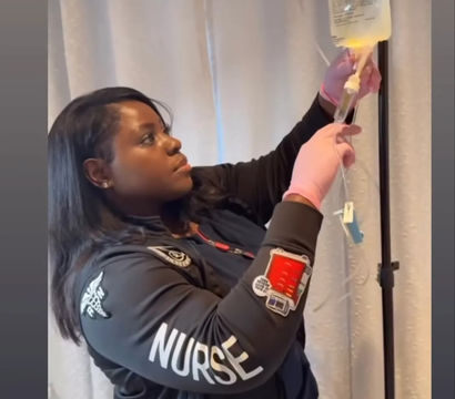 Nurse wearing pink gloves adjusts an IV drip bag on a pole in a patient room, jacket sleeve clearly labeled "NURSE".