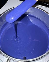 Glossy, glittery royal blue paint pouring from a spatula into a metal paint can, smooth vibrant finish.