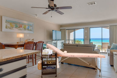 Bright beachfront condo living room with tile floors, sofa and TV facing sliding glass doors to a turquoise ocean balcony; dining table, ceiling fan, and a pink ironing board with a utility cart in the foreground.