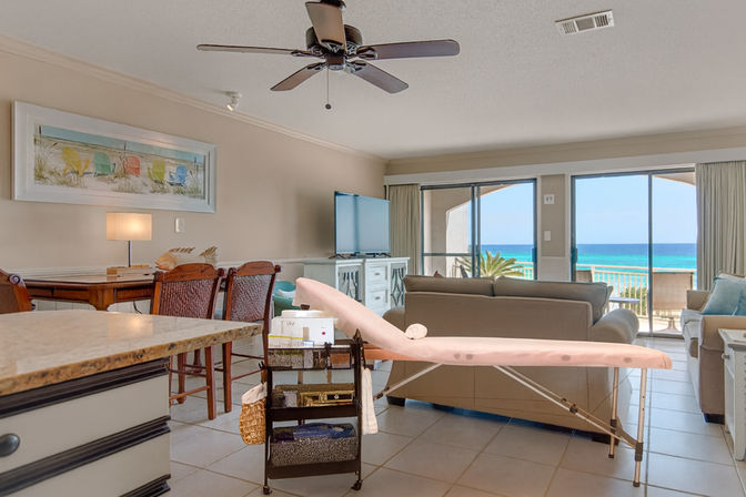 Bright beachfront condo living room with tile floors, sofa and TV facing sliding glass doors to a turquoise ocean balcony; dining table, ceiling fan, and a pink ironing board with a utility cart in the foreground.