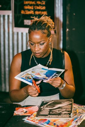 Focused person with dreadlocked hair and glasses cutting a magazine with red scissors at a table of colorful collage materials in an indoor arts-and-crafts workshop