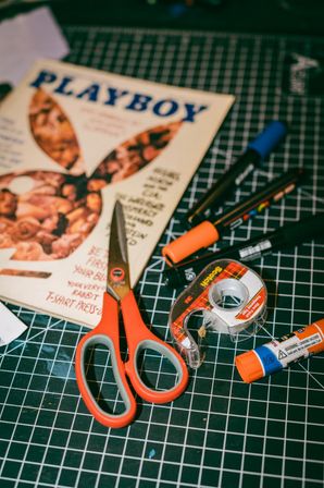 DIY craft desk on a black grid cutting mat with orange-handled scissors, clear tape dispenser, orange glue stick, multi-color markers and a vintage magazine cover.
