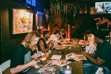 Group of people cutting magazines and making collages at a long table in a cozy, dimly lit art gallery workshop with framed artwork, hanging greenery and warm accent lighting.