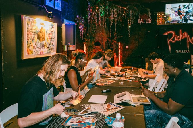 Group of people cutting magazines and making collages at a long table in a cozy, dimly lit art gallery workshop with framed artwork, hanging greenery and warm accent lighting.