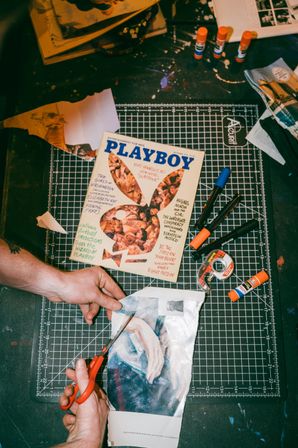 Hands cutting a printed image on a black grid cutting mat, surrounded by a vintage Playboy magazine, scissors, glue sticks, markers and tape in a messy tabletop collage craft workspace.