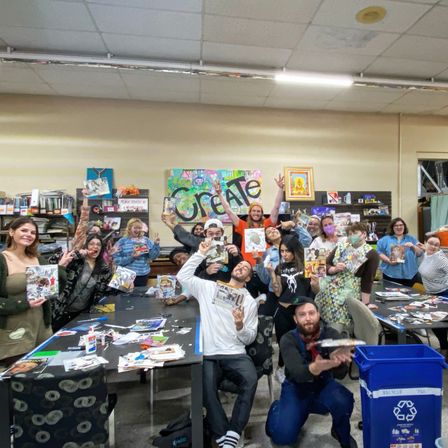 Lively community art workshop in a studio: a large group of people posing with handmade collages at long tables covered in glue, scissors and paper, colorful 'CREATE' mural on the back wall and a recycling bin in the foreground