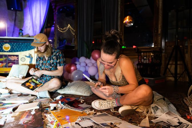 Two people sitting on a rug in an indoor art studio, surrounded by magazines and torn paper; a woman in a leopard tank and sneakers cuts paper with scissors while a person in a patterned shirt and cap sorts materials, with pastel balloons and string lights in the background suggesting a casual craft party.