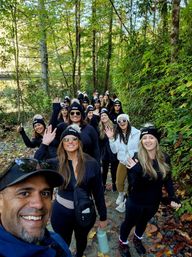 Smiling group hike on a forest trail in fall — people waving at the camera wearing matching black pom‑pom beanies and sunglasses, autumn leaves on the path and dense green trees with a glimpse of water beyond.