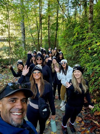 Smiling group hike on a forest trail in fall — people waving at the camera wearing matching black pom‑pom beanies and sunglasses, autumn leaves on the path and dense green trees with a glimpse of water beyond.