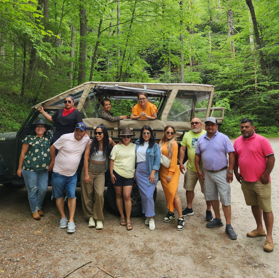 Dozen adults posing beside an open-top safari jeep in a lush green forest clearing, wearing casual summer outfits and sunglasses during an outdoor adventure tour.