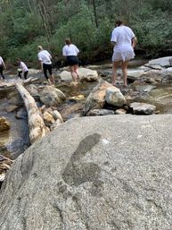 Wet barefoot footprint on a boulder in the foreground, group of people wading across a rocky forest stream on an outdoor hike