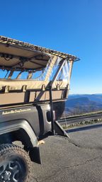 Open-air tour Jeep with tan canvas canopy and clear vinyl windows parked at an Asheville mountain overlook, showing rugged tire, asphalt pullout, and layered blue ridges under a clear sky.