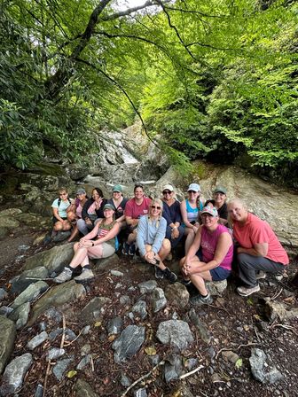 Smiling hiking group resting on a rocky streambed beneath a small waterfall in a lush green forest canopy.