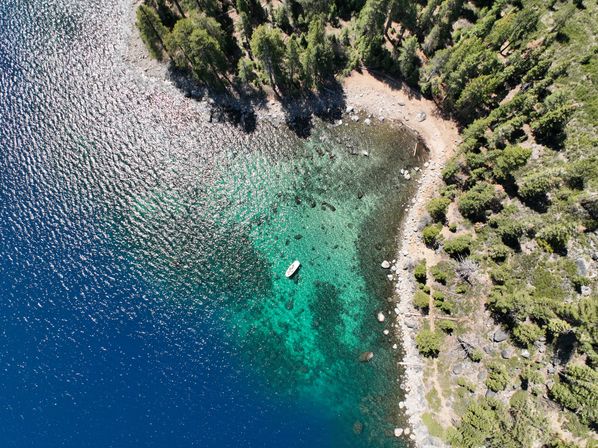 Aerial view of a clear turquoise lake cove with a small white boat floating near a rocky, pine-lined shoreline and deep blue water.