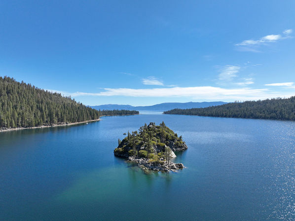 Aerial view of a tiny pine‑covered rocky island in a crystal‑blue mountain lake, framed by forested shorelines and distant ridgelines under a sunny sky.