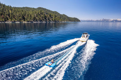 Speedboat pulling a bright blue towable tube with a rider across a deep-blue alpine lake, pine-forested shore and distant snow-capped mountains.