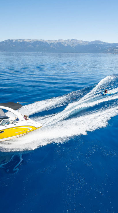 Yellow speedboat towing a waterskier across a glassy deep-blue mountain lake, carving a foamy white wake under a clear sky.