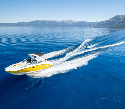 Yellow speedboat cruising across a glassy blue mountain lake, leaving a white wake while towing a person on an inflatable tube under a clear sky.