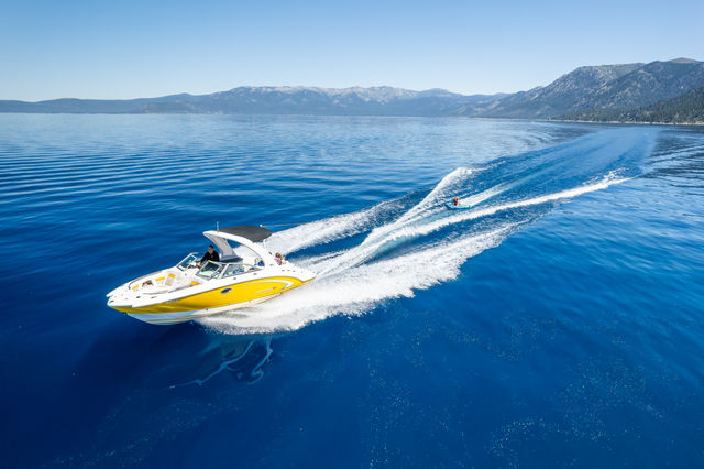 Yellow speedboat cruising across a glassy blue mountain lake, leaving a white wake while towing a person on an inflatable tube under a clear sky.