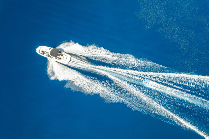 Aerial view of a white speedboat towing a colorful inflatable tube across deep blue open sea, leaving foamy twin wake trails in a sunny water-sports scene