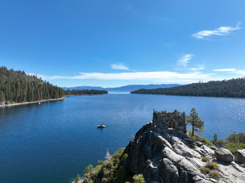 Scenic mountain lake with deep blue water, pine‑covered shores, a small boat on the water, and a stone lookout perched on a rocky cliff under a clear blue sky.