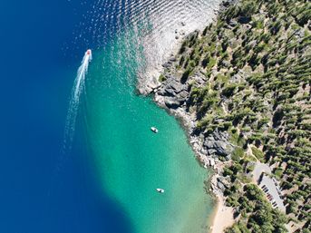 Aerial view of a turquoise mountain lake cove with a speedboat leaving a white wake, two anchored boats, rocky pine-lined shore and a small sandy beach.