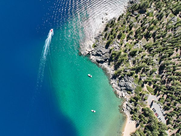 Aerial view of a turquoise mountain lake cove with a speedboat leaving a white wake, two anchored boats, rocky pine-lined shore and a small sandy beach.