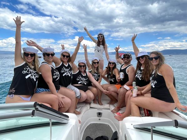 Group of people in matching 'Lake Life' tank tops and sailor hats cheering with raised arms and drinks aboard a motorboat on a sunny mountain lake under a dramatic blue sky.