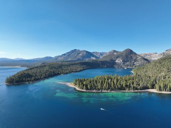 Aerial view of an alpine lake with clear blue and turquoise shallows, a tree-covered peninsula and pine-clad mountains under a bright sky, a small motorboat leaving a wake