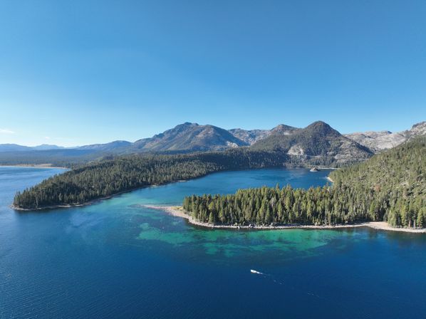 Aerial view of an alpine lake with clear blue and turquoise shallows, a tree-covered peninsula and pine-clad mountains under a bright sky, a small motorboat leaving a wake