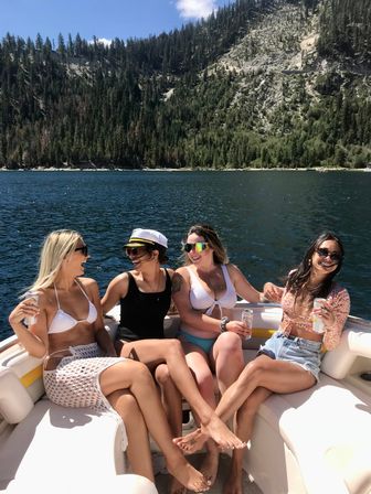 Four friends laughing on a motorboat in swimsuits, holding canned drinks on a sunny summer day cruising a deep-blue mountain lake with pine-covered cliffs in the background.