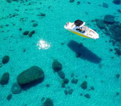 Aerial view of a white-and-yellow speedboat anchored in crystal-clear turquoise water, sun-speckled surface revealing scattered submerged rocks and the boat's dark shadow on the sandy seabed.